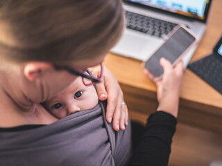 Mutter mit Handy in der Hand und Laptop auf dem Tisch sieht ihr Baby in der Trage an